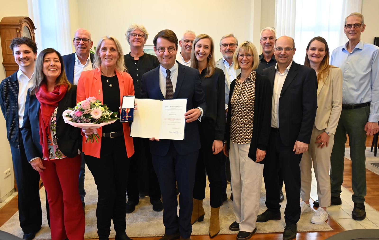 Eine Gruppe von 14 Personen lächelt, eine Person hält ein Dokument und andere Blumen.