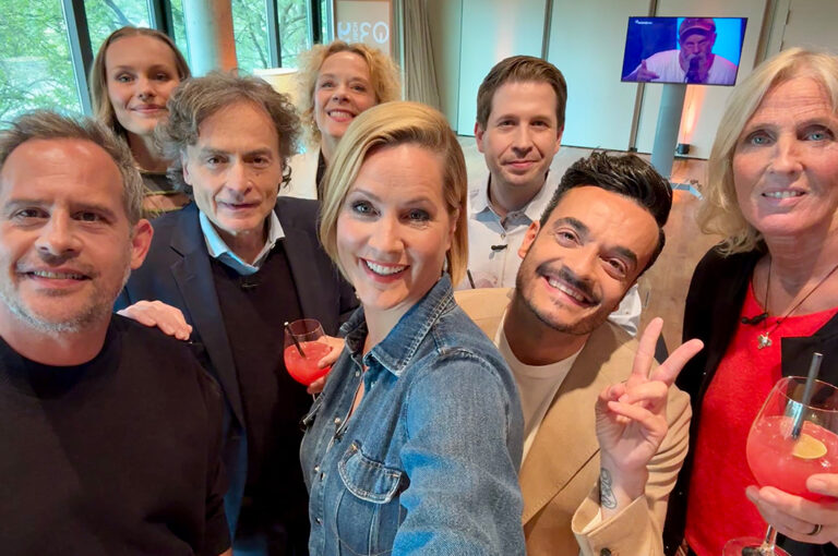 Acht lächelnde Personen posieren für ein Gruppenfoto bei einem Event im Innenraum