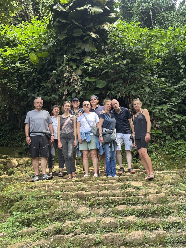 Gruppe von acht Personen vor grüner Laubwand, lächeln, lässig gekleidet. Treppen aus Stein.