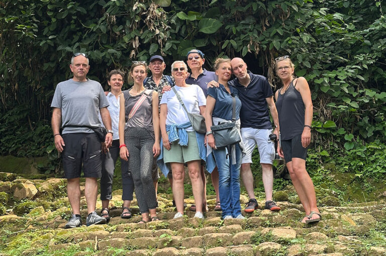 Gruppe von neun Personen lächelt entspannt vor üppigem, grünem Hintergrund.