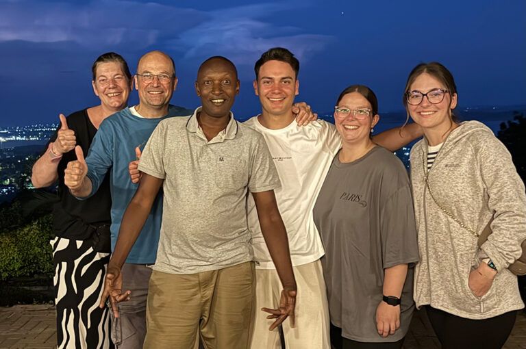 Eine Gruppe von sechs Personen lächelt draußen bei Abendhimmel in die Kamera.