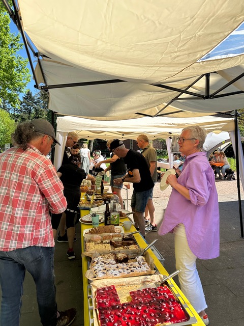 Eine Gruppe Menschen bei einem Marktstand, Frau in lila Hemd lacht, viele Backwaren ausgestellt.