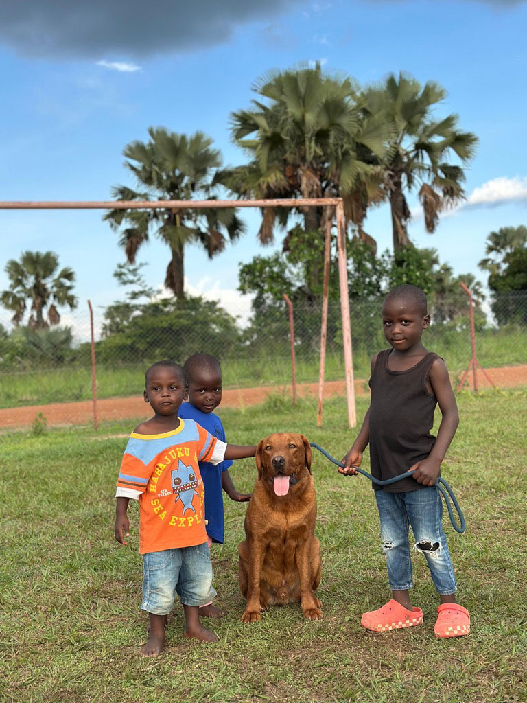 Drei Kinder mit neutralem Gesichtsausdruck stehen mit einem braunen Hund auf einem grünen Feld.