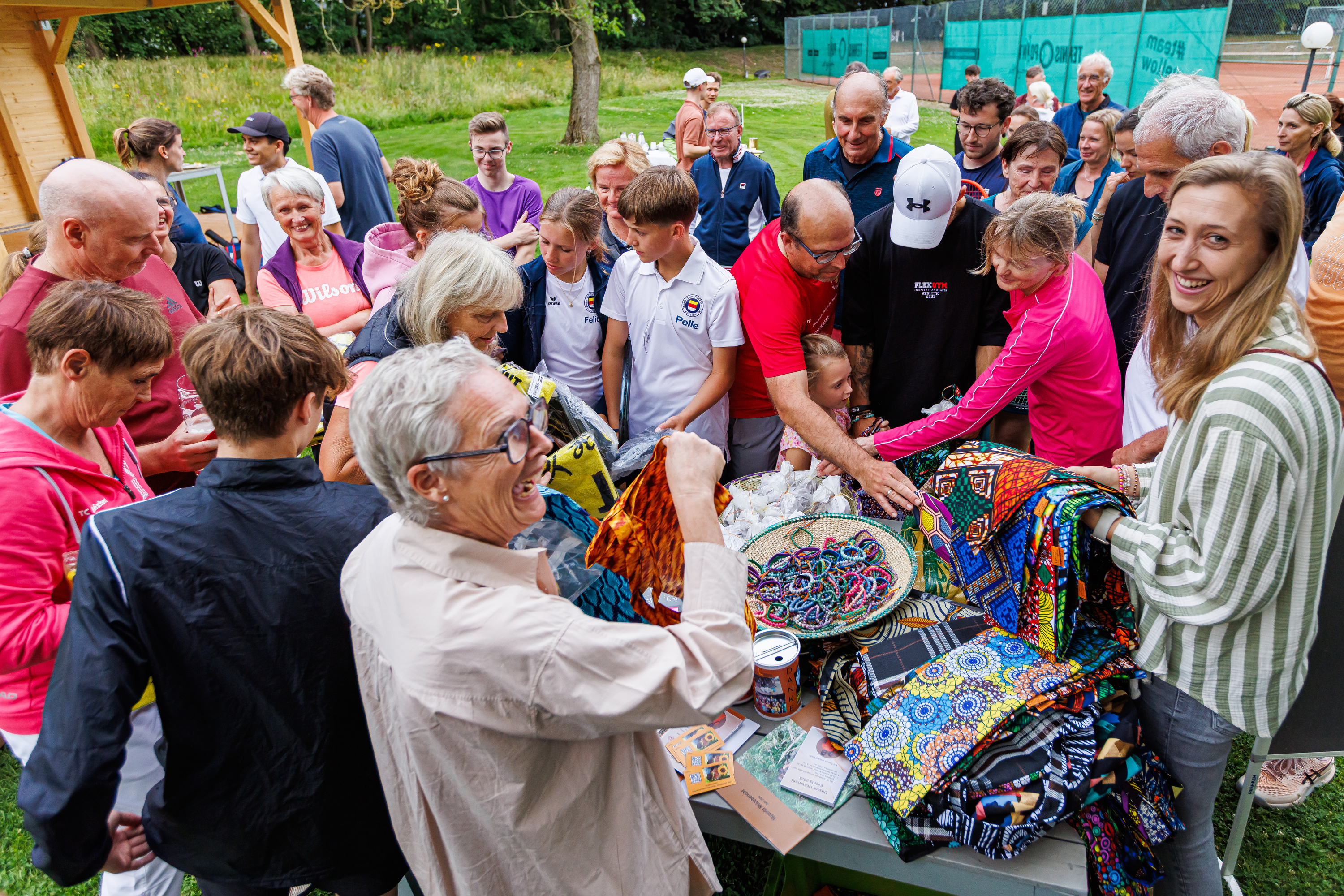 Menschen bei einem Outdoor-Event, fröhlich, bunt gekleidet, betrachten Tücher auf einem Tisch.
