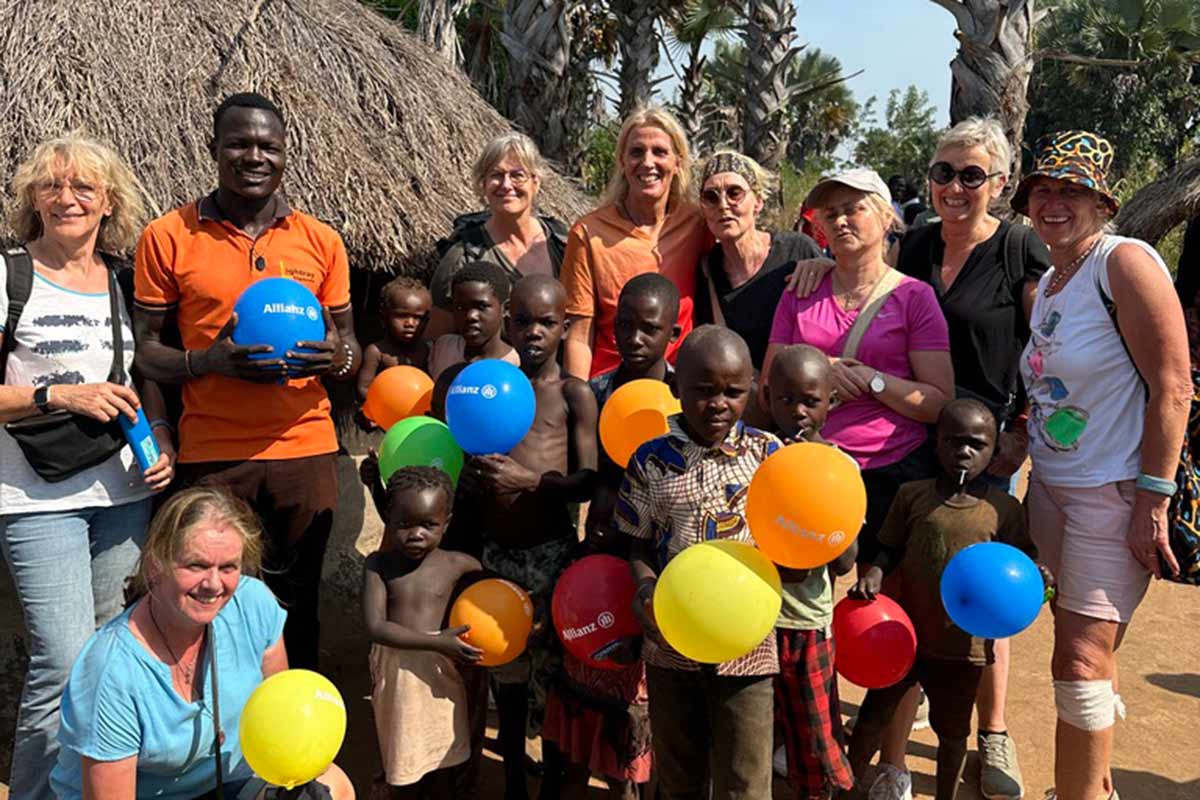 Eine Gruppe fröhlicher Menschen hält bunte Ballons. Hintergrund: strohgedecktes Gebäude.