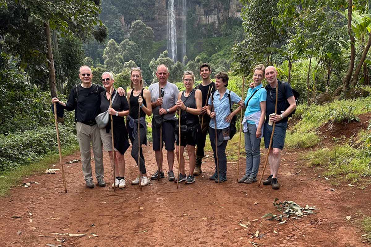 Gruppe von Wanderen lächelt vor einem Wasserfall; Bäume und roter Erdboden im Hintergrund.