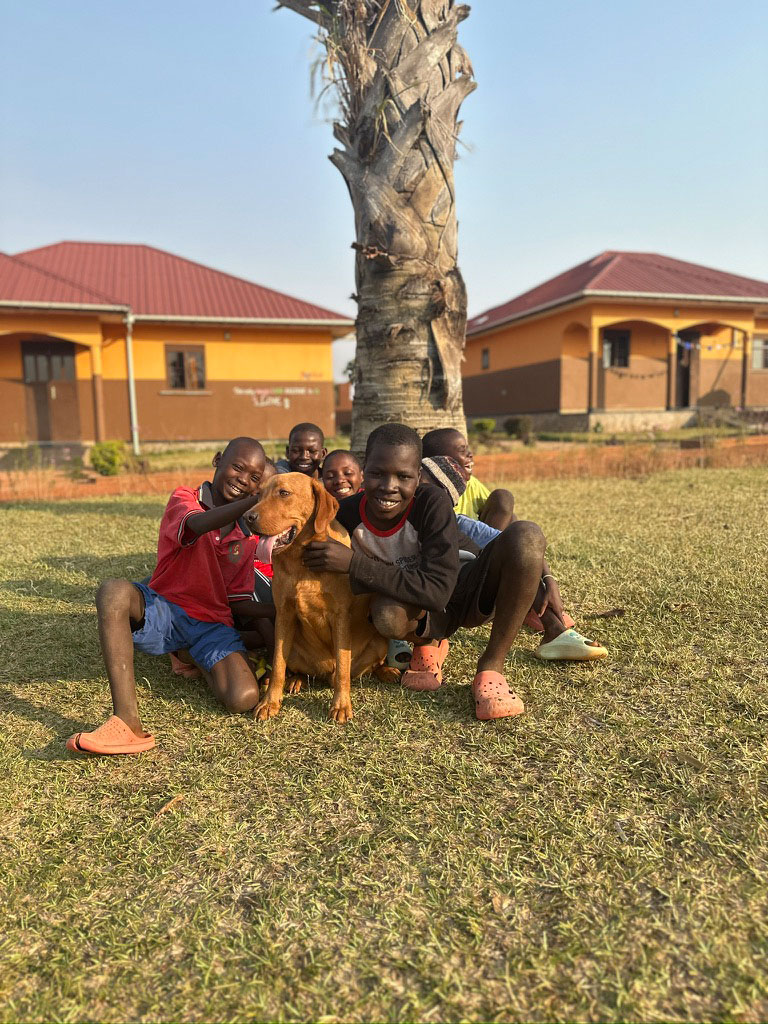 Gruppe von Kindern mit Hund im Gras; lächelnd, farbenfrohe Kleidung, rotes Haus im Hintergrund.