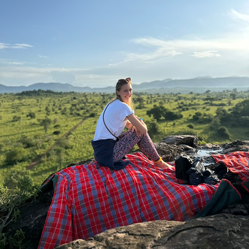 Eine lächelnde Frau sitzt auf einer karierten Decke mit Blick auf eine weite grüne Landschaft.