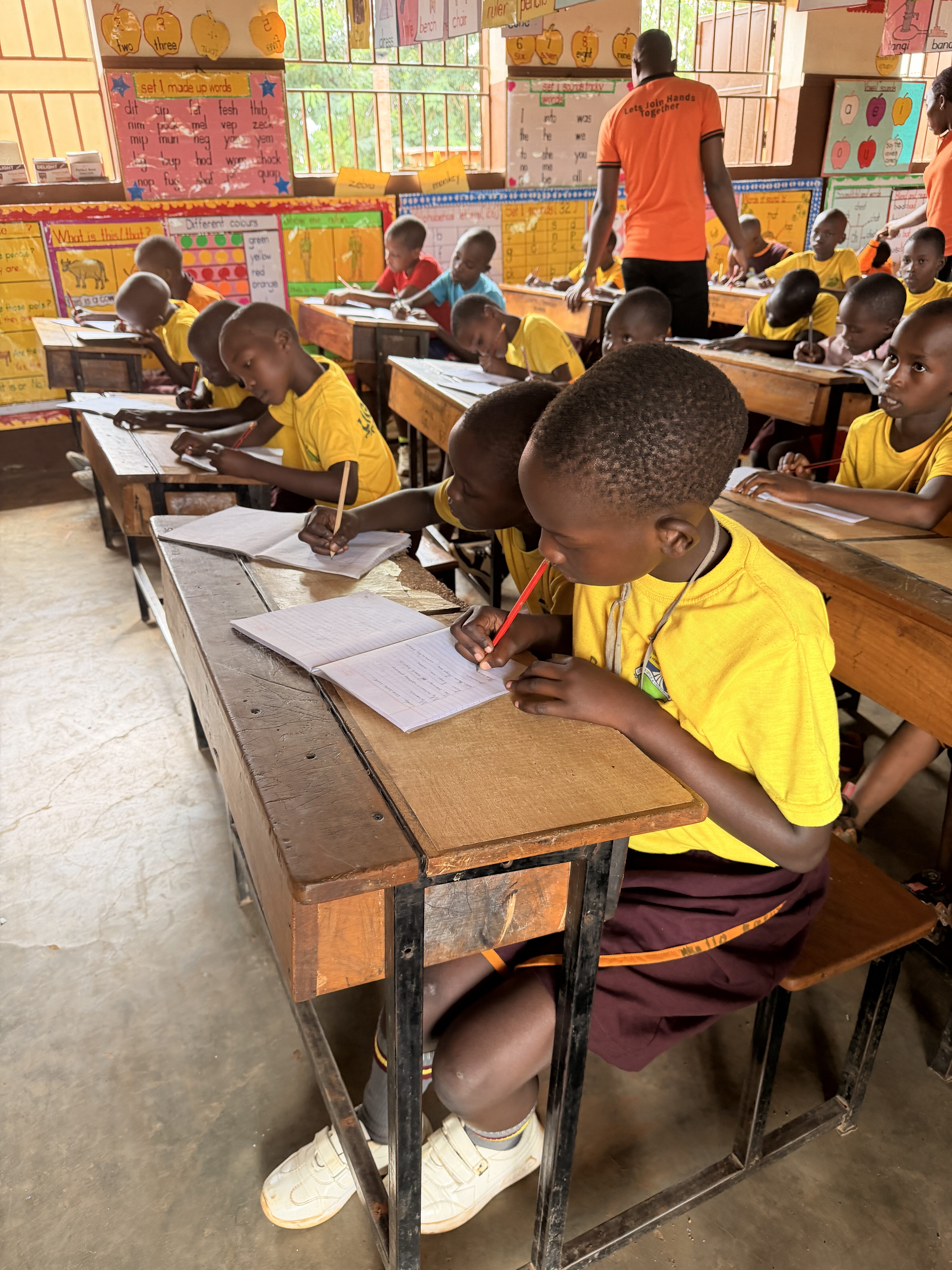 Konzentrierte Schulkinder in gelben Shirts schreiben in bunter Klasse, Lehrer in Orange