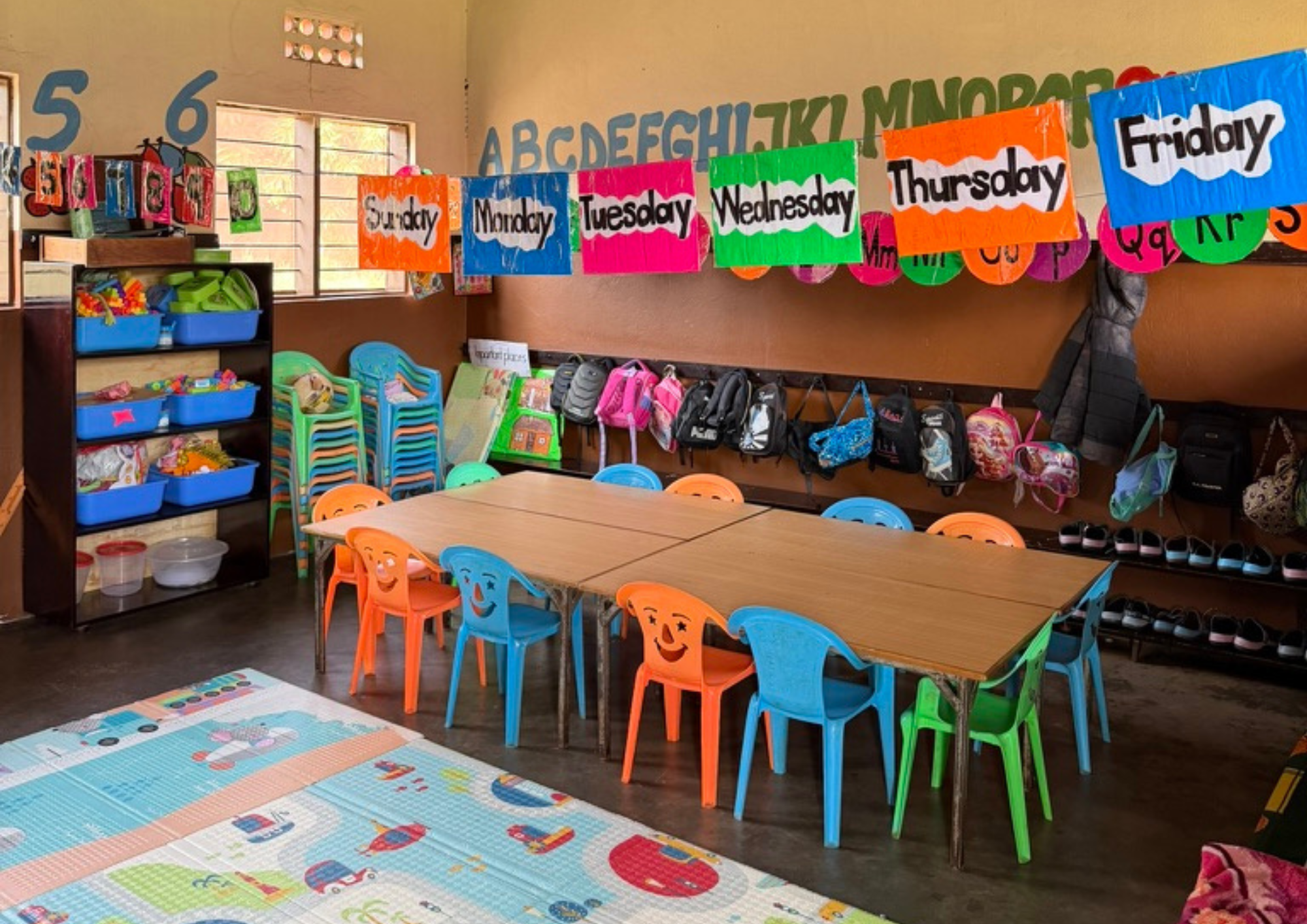 Bunter Kindergartenraum mit Tischen Stuehlen Rucksaecken und Wochentagsschildern
