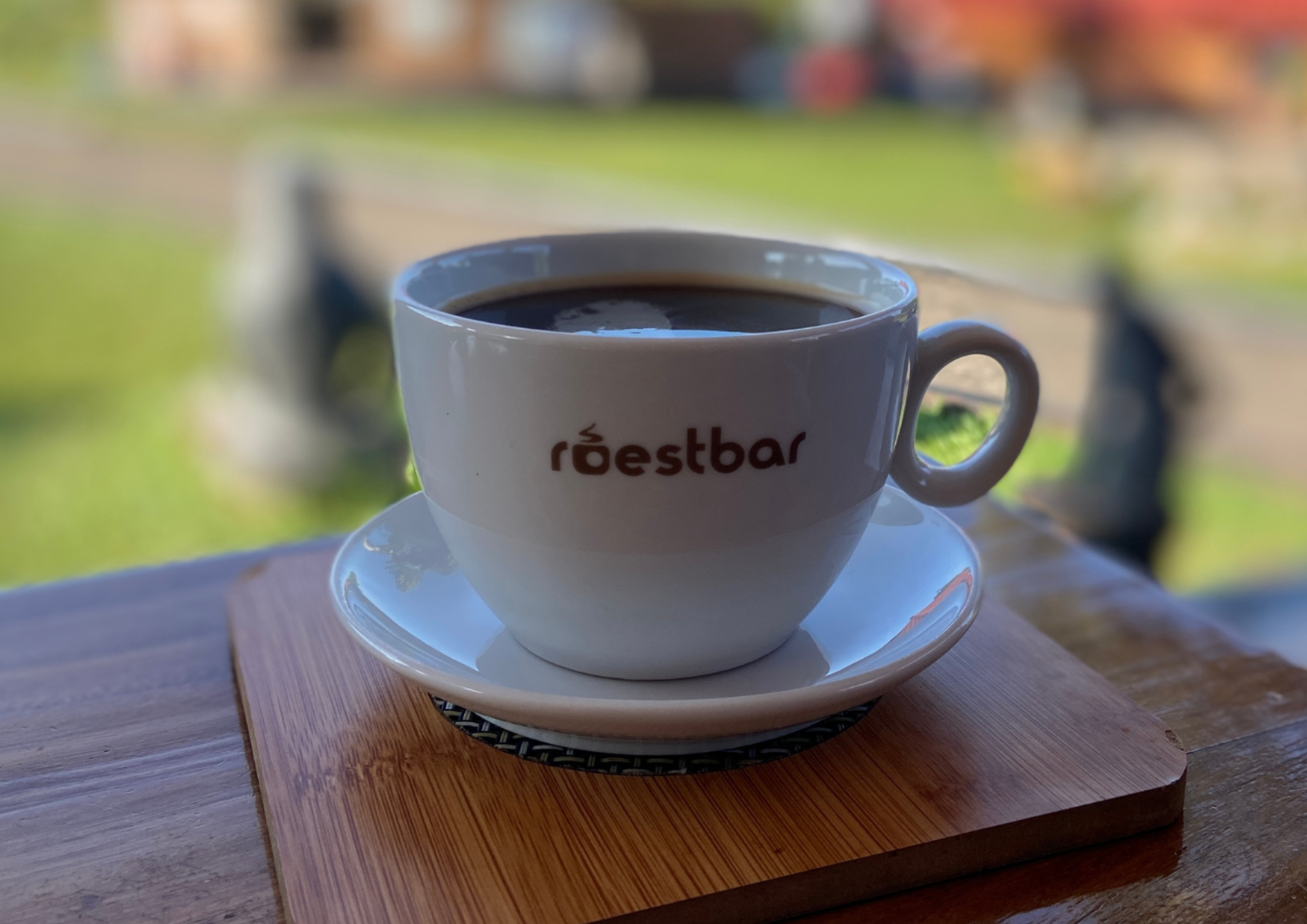 Weisse roestbar Tasse mit schwarzem Kaffee auf Holzbrett vor gruenem Hintergrund