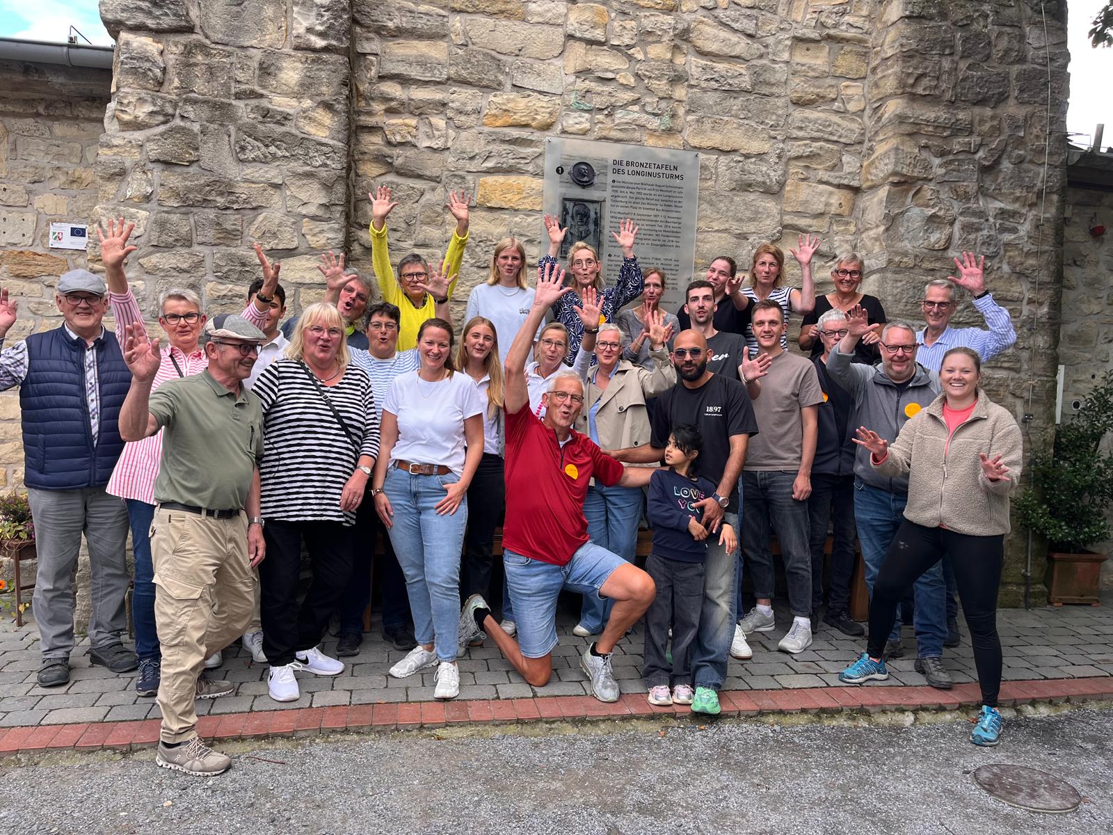 Eine froehliche Gruppe winkt vor einer Steinmauer mit Bronzetafeln